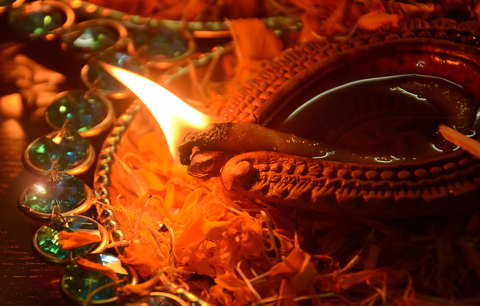 Traditional Diwali diya, set on a small bejeweled dish, covered with flower petals. Inside the diya, a large wick is burning. The entire image is cast in warm orange light from the flame of the wick. 