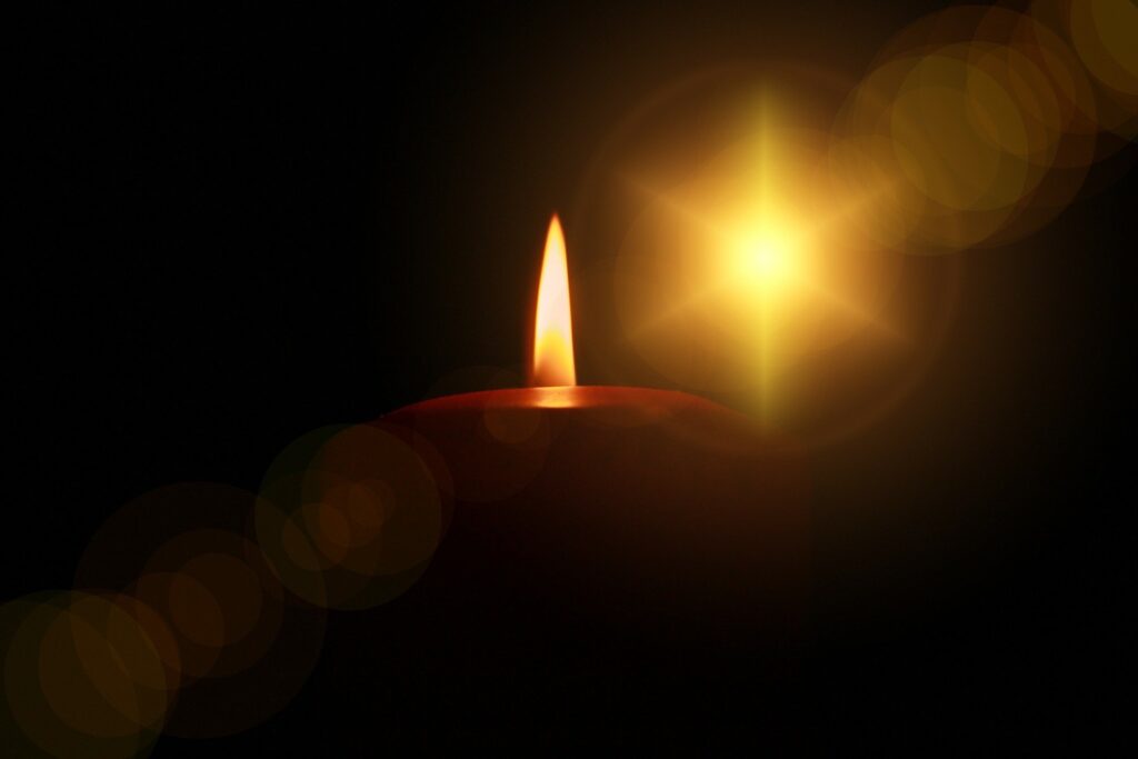 Just the top portion of a single, red, lit candle is highlighted against a dark background. Diagonally across the image is the diffused light of that candle.